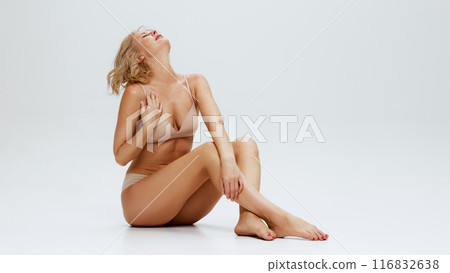 Blonde woman in nude lingerie sits on floor, her head tilted back in moment of relaxation and joy against white studio background. 116832638