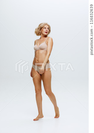 Middle-aged woman with blonde hair wears minimalistic nude lingerie highlights her slim figure against white studio background. 116832639