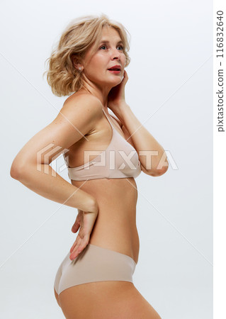 Photo of elegant woman in nude underwear poses gracefully, her hand resting on her hip against white studio background. 116832640