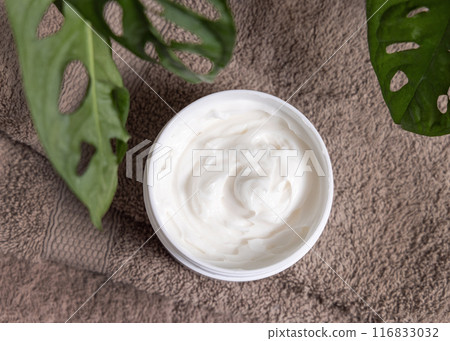 Opened cream jar on brown towel near green plant top view, everyday facial care 116833032