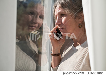 Serious aged latin female pensioner stand by picture window at home hold phone 116833084