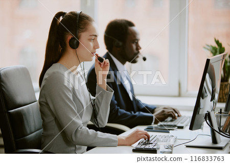 Young Caucasian operator speaking with client via wired headphone while his African American coworker searching information in program in blurred background 116833763