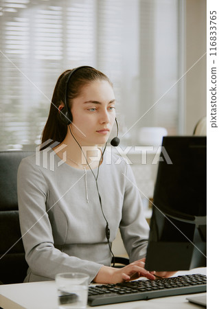Tired young female call center operator looking at monitor and typing on keyboard while wearing wired headphones with microphone Tired young female call center operator looking at monitor and typing on keyboard while wearing wired headphones with microphone 116833765