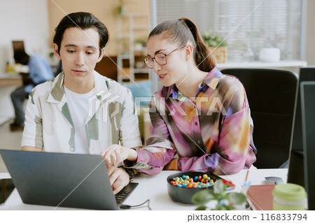 Two young colleagues discussing final code while sitting at white table with electronic devices and snacks on top in modern comfort office Two young colleagues discussing final code while sitting at white table with electronic devices and snacks on top in modern comfort office 116833794