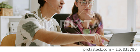 Website header shot of two coworkers sitting at table desk and looking at laptop screen in office 116833796