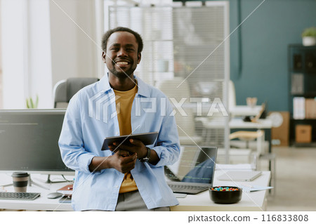 Young African American IT specialist holding silver touchpad and smiling widely while leaning on table in modern and comfortable office of high-end IT company Young African American IT specialist holding silver touchpad and smiling widely while leaning on table in modern and comfortable office of high-end IT company 116833808