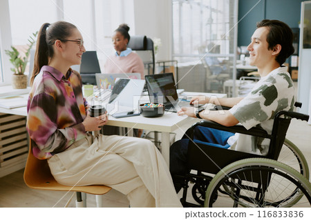 Young Caucasian female colleague holding cup of coffee and chatting lovely with her Caucasian male colleague with disability 116833836