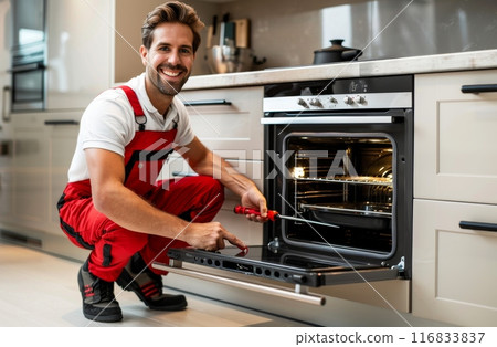 Smiling repairman in red overalls fixing an oven in the kitchen 116833837