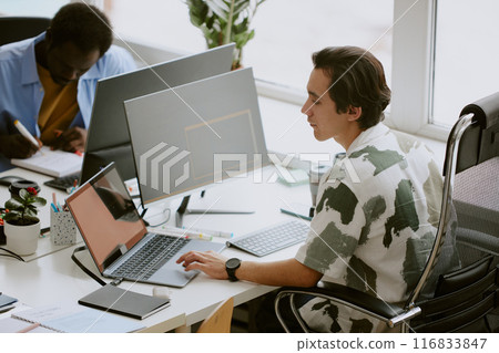 Multitask Caucasian programmer using gray laptop to search information while using computer to write code, his African American coworker writing report in blurred background 116833847