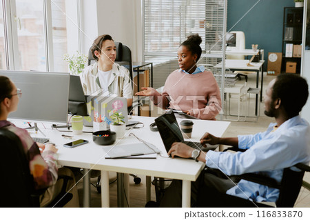 Biracial colleagues discussing next goals while sitting at white table in modern IT company office equipped with electronic devices 116833870