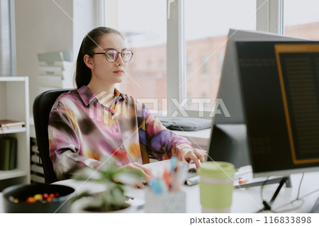 Young Caucasian female programmer dressed in spotted blouse sitting at work desk while typing on keyboard and looking directly at computer screen Young Caucasian female programmer dressed in spotted blouse sitting at work desk while typing on keyboard and looking directly at computer screen 116833898