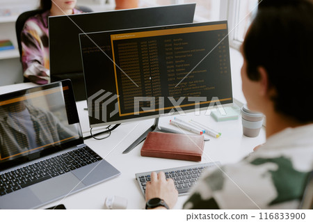 Over shoulder shot of male programmer surrounded by computers looking at screen with launched app while coding, his coworker sitting in front of him 116833900