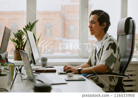 Profile shot of young programmer sitting at wooden work table while coding via computer in light office 116833908