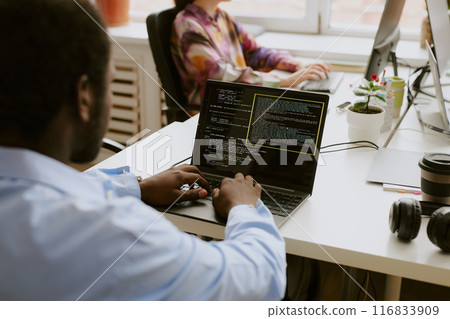 Over shoulder shot of African American man sitting at white wooden table and writing codes while his coworker typing on keyboard in blurred background 116833909