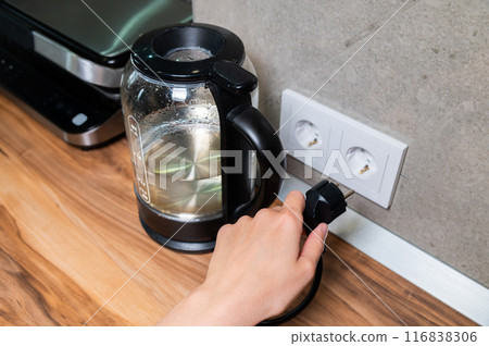 glass modern electric kettle stands on the table against the background of an outlet. Boiling water. Electricity consumption by household electrical appliances glass modern electric kettle stands on the table against the background of an outlet. Boiling water. Electricity consumption by household electrical appliances 116838306