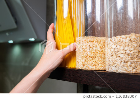 grains and pasta are stored in a jar on a shelf in a cabinet in the kitchen, and a woman's hand takes a plastic container with spaghetti for cooking 116838307