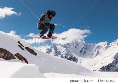 Jumping snowboarder holds one hand on a snowboard against the background of a blue sky and large snow-capped mountains. Performing a stunt in flight 116838308