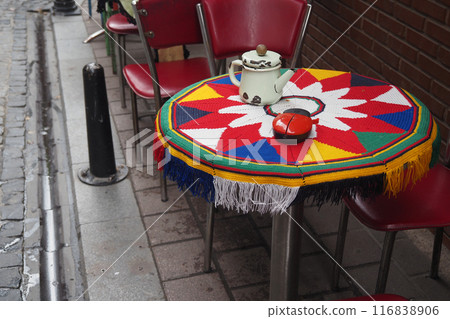 Colorful outdoor cafe scene with a teapot on a patterned table, surrounded by red chairs 116838906