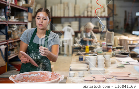 Young female worker in pottery factory lowers fired clay plates and cups into container with glaze 116841169