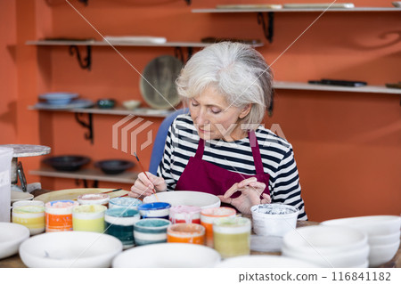 Mature craftswoman painting clay plates acrylic paints in art studio 116841182
