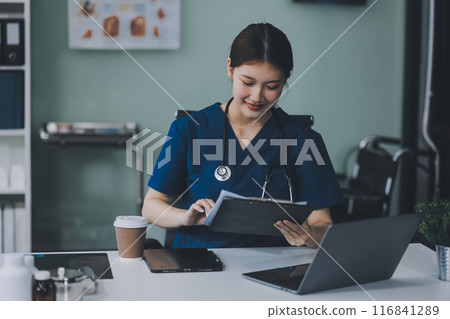 Female doctor writing in a notebook and holding a medicine bottle works at a computer while there were many medicine bottles on the table while sitting at a work desk Female doctor writing in a notebook and holding a medicine bottle works at a computer while there were many medicine bottles on the table while sitting at a work desk 116841289