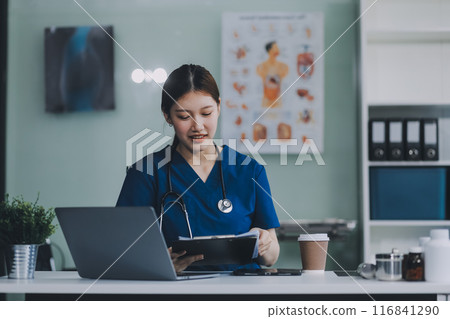 Female doctor writing in a notebook and holding a medicine bottle works at a computer while there were many medicine bottles on the table while sitting at a work desk Female doctor writing in a notebook and holding a medicine bottle works at a computer while there were many medicine bottles on the table while sitting at a work desk 116841290