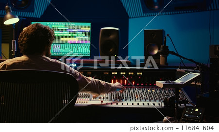 Tracking engineer processing and mixing sound on audio console with moving faders and colored meters. Music producer working on a new album, creating a hit song in studio. Camera B. 116841604