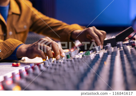 African american sound engineer twisting knobs to edit music in studio, operating control panel board desk with motorized faders and buttons. Skilled producer creating records with gear. Close up. African american sound engineer twisting knobs to edit music in studio, operating control panel board desk with motorized faders and buttons. Skilled producer creating records with gear. Close up. 116841617