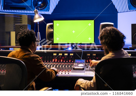 Team of musicians editing music with greenscreen on computer in professional recording studio, using audio mixing console with knobs and faders in control room. Artist and producer compose tracks. Team of musicians editing music with greenscreen on computer in professional recording studio, using audio mixing console with knobs and faders in control room. Artist and producer compose tracks. 116841660