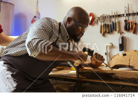 BIPOC man in carpentry shop wearing protection equipment while carving wood to prevent workplace accidents. Cabinetmaker equipped with safety goggles while using chisel and hammer to avoid injury BIPOC man in carpentry shop wearing protection equipment while carving wood to prevent workplace accidents. Cabinetmaker equipped with safety goggles while using chisel and hammer to avoid injury 116841714