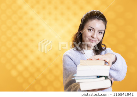 Woman holding stack of books, pointing it towards camera, isolated over yellow studio background. Portrait of smiling bookworm lady presenting stack of novels, urging people to read 116841941