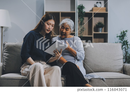 Happy grownup daughter showing content on tablet to mature mother. Two family generations women with digital computer gadget resting on couch together, watching videos, making video call 116843071
