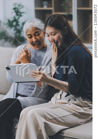 Happy grownup daughter showing content on tablet to mature mother. Two family generations women with digital computer gadget resting on couch together, watching videos, making video call 116843072