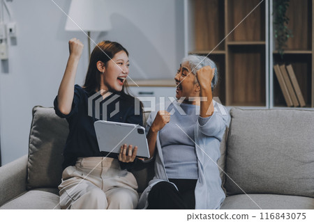 Happy grownup daughter showing content on tablet to mature mother. Two family generations women with digital computer gadget resting on couch together, watching videos, making video call 116843075