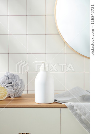 Close up shot photography at an unlabeled shower gel bottle in white color, which placed above of wooden cabinet. On both side, displaying some bathroom accessories such as towel and bath sponge 116843721