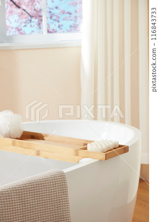 Bathroom scene with japan style, a white large bathtub placed by the window with a wooden tray above, which contains soap and bath sponge. High angle view, blank space for designing 116843733