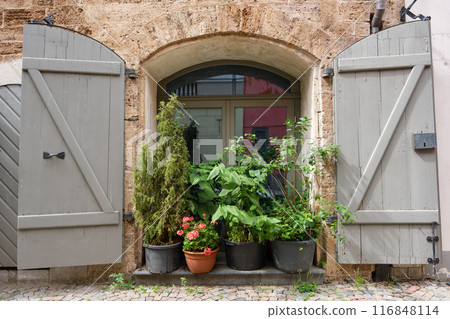 Vintage window with wooden shutters or front door on a building facade decorated with plants and flowers in pots. Old town of Riga, Latvia. Vintage window with wooden shutters or front door on a building facade decorated with plants and flowers in pots. Old town of Riga, Latvia. 116848114