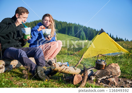 Man and woman enjoying meals from food pouches. Tourists sit on log near campfire. Behind them, yellow tent pitched on lush green hillside, with trees in background. 116849392