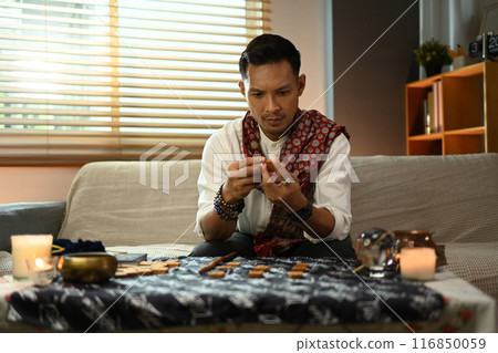 Male fortune teller in traditional attire forecasting with wooden runes. Divination concept Male fortune teller in traditional attire forecasting with wooden runes. Divination concept 116850059