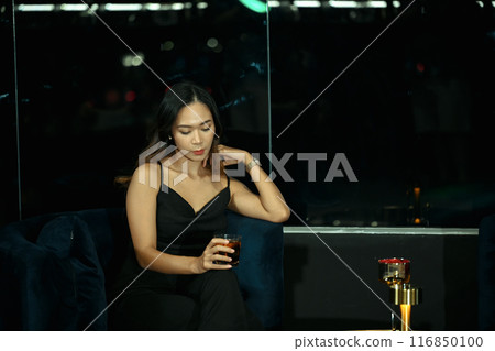 Elegant young woman in evening dress holding a drink sitting alone in a dimly lounge. Nightlife and luxury lifestyle concept Elegant young woman in evening dress holding a drink sitting alone in a dimly lounge. Nightlife and luxury lifestyle concept 116850100