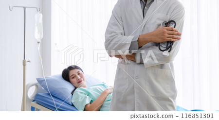 Male doctor standing confidently by patient bedside with stethoscope in hand at hospital ward room. Asian young woman resting comfortably on hospital bed. 116851301