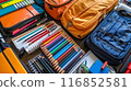 Professional photograph of a school backpack brimming with supplies. The backpack, positioned upright, is slightly open, showcasing various school essentials. Visible items include spiral notebooks, 116852581