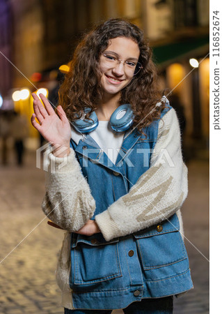 Teenage girl waving hands gesturing hi hello greeting or goodbye on urban city street at night Teenage girl waving hands gesturing hi hello greeting or goodbye on urban city street at night 116852674