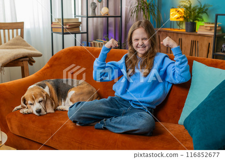 Cheerful young child girl celebrating victory winning the lottery at home beside beagle dog on sofa Cheerful young child girl celebrating victory winning the lottery at home beside beagle dog on sofa 116852677