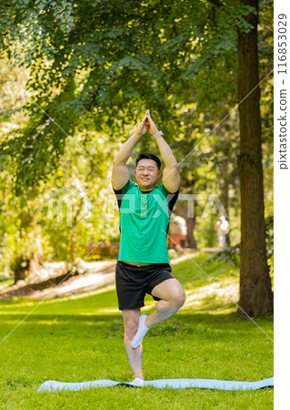 Calm muscular Asian Japanese sportsman guy balances yogi training on one leg on mat in summer park 116853029