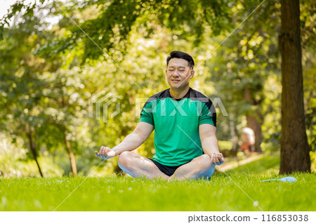 Young Asian man sitting on mat in lotus position, relaxing, practicing yoga in park on summer day Young Asian man sitting on mat in lotus position, relaxing, practicing yoga in park on summer day 116853038