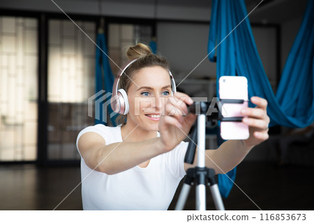 Young beautiful woman in headphones smiling at smartphone camera, she is teaching online yoga class Young beautiful woman in headphones smiling at smartphone camera, she is teaching online yoga class 116853673