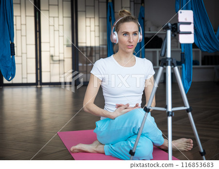 Young slim blonde woman in sportswear in yoga hall with headphones in front of smartphone on tripod Young slim blonde woman in sportswear in yoga hall with headphones in front of smartphone on tripod 116853683
