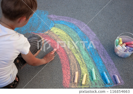 A boy draws a rainbow with multi-colored chalk on the asphalt. Drawing multicolored chalk stick, back to school background. Top view. A boy draws a rainbow with multi-colored chalk on the asphalt. Drawing multicolored chalk stick, back to school background. Top view. 116854663