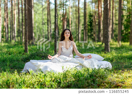 Young woman is sitting in the forest on a white bed Young woman is sitting in the forest on a white bed 116855245